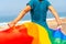 Unrecognizable lesbian person in a blue dress and white hat waving the LGBTQ flag by the sea