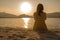 Unrecognizable lonely pensive woman sitting on the sand of seashore looking at the setting sun with light reflection in the water