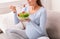 Unrecognizable Pregnant Girl Eating Salad From Bowl Sitting On Sofa