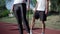 Unrecognizable slim woman in black leggings stretching on sunny running track with man warming up at the background