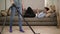 Unrecognizable woman in jeans vacuuming parquet floor, and her husband plays on smartphone on couch. Front view, slow