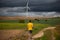 Unrecognizable young adult man walking through a farm field with sustainable windmill at the background. Epic dark sky.