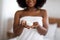 Unrecognizable young African American female in bath towel holding jar of face or body cream at home, closeup