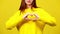 Unrecognizable young woman making heart gesture standing at yellow background smiling. Slim young Caucasian lady