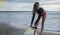 Unrecognizble barefoot woman has fixed legrope, stands on sand near surfboard, protects herself from crashing into shore lines,