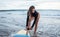 Unrecognizble barefoot woman has fixed legrope, stands on sand near surfboard, protects herself from crashing into shore lines,