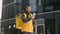 Unshaven courier with yellow backpack on his back in uniform walks down street in winter among tall glass buildings