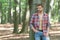 Unshaven lumbersexual man in unbuttoned lumberjack shirt forest background, copy space