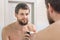An unshaven man applies shaving foam on the palm of his hand, and looks in the mirror in the bathroom