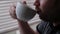 An unshaven man drinks tea near a window closed with blinds