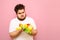 Upset bearded fat man in white t-shirt stands on a pink background with fruits in his hands. Overweight man on a diet eats fruits