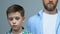 Upset boy looking into camera, father standing on background, assault at home