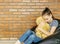 Upset caucasian teen girl sitting in black bean bag chair hug big brown teddy bear toy against brick wall. Casual outfit. Sadness