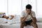 Upset depressed young man sitting on the edge of bed against his wife lying on the bed. Relationship difficulties