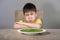 Upset and disgusted Asian kid sitting on table refusing to eat spinach thick soup looking unhappy rejecting vegetarian food in