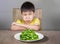 Upset and disgusted hispanic kid sitting on table in front of spinach plate unhappy rejecting the fresh food finding it disgusting