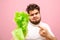 Upset fat curly man points his finger at salad leaves in his hand and looks into the camera with an evil face on a pink background