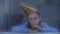 Upset girl in birthday hat blowing party horn on rainy day, celebrating alone