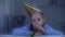 Upset girl in birthday hat blowing party horn on rainy day, celebrating alone