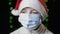 Upset girl in red Santa Claus hat, blue medical protective mask looks at camera