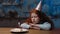 Upset little girl with long red curly hair sitting at table with head down on hands. Lonely kid in birthday hat looking