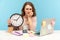 Upset nervous woman employee sitting at workplace office, all covered with sticky notes and holding big clock