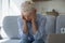 Upset older woman holding head with hands seated on sofa