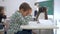 Upset schoolboy sits at a desk in classroom on background of classmates and female teacher near blackboard