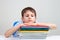 Upset schoolboy sitting at desk with pile of school books and notebooks