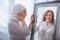 upset sick mature woman in kerchief looking at smiling reflection in mirror