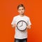 Upset teen boy holding big clock, orange studio background