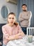 Upset woman sitting at table on background of displeased husband