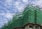 Upward view of precast building, scaffolding on each story cover by green net in construction work, under white fluffy cloud