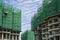 Upward view of precast building, scaffolding on each story cover by green net in construction work, under white fluffy cloud