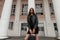 Urban attractive young woman with brown hair with in a leather stylish jacket in a black dress with a black fashionable bag