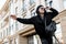 Urban cheerful young man hitchhiker in a cap in trendy sunglasses with a stylish backpack in a black sweatshirt is standing