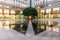 Urban space fountain area with big green tree in the corner with people. This area amid modern office buildings left and right.