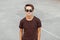 Urban trendy young hipster man in stylish brown t-shirt with hairstyle in trendy black sunglasses stands at the stadium.