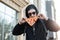 Urban young hipster man in trendy sunglasses in a black fashionable sweatshirt with a hood in a cap holds a slice of pizza