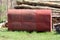 Used old red metal oil storage tank surrounded with uncut grass in front of firewood and wooden logs