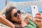 Vacation woman using smart phone taking smartphone selfie. Girl relaxing on beach hammock sun tanning in sunglasses smiling using