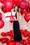 Valentine`s day concept - middle aged beautiful woman in long black dress with big gift box and red balloons