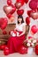 Valentine`s day concept - young woman sitting on vintage sofa and opening gift box with red heart-shaped balloons