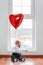 Valentine`s day. Little baby boy looks out the window, with a red ball in the form of a heart. Vertical