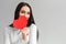 Valentine`s Day. Woman standing isolated on gray hiding face behind heart card looking camera playful close-up