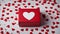 valentines day card A red gift box with a white heart-shaped cutout on it. The box is surrounded with confetti hearts