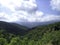 Valley of hills covered with low green forest. In the distance, the silhouettes of high mountains turn blue. Clear blue sky with