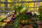 A variety of autumn flowers in ceramic pots and vegetables in baskets stand on a shelf covered with burlap and straw.