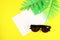 Various tropical leaves with a white sheet of paper sunglasses on a yellow background, top view