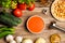 Vegetables needed to make a gazpacho around a bowl filled with gazpacho and chunks of toasted bread, on a rustic wooden table, top